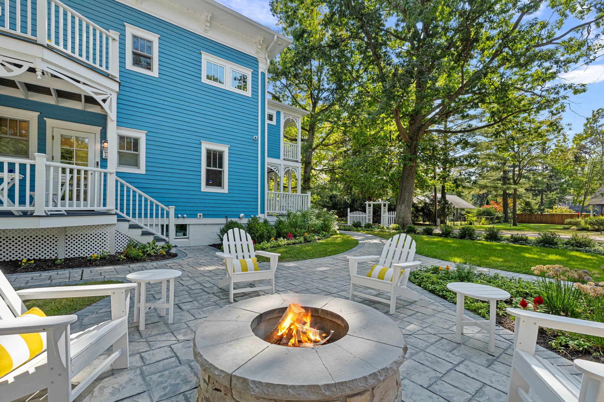 A circular stone fire pit with a bright flame is centered on a grey paved patio. Several white Adirondack chairs with yellow and white striped pillows are arranged around the fire, set against the backdrop of a vibrant blue house with white trim and a lush green lawn shaded by large trees.
