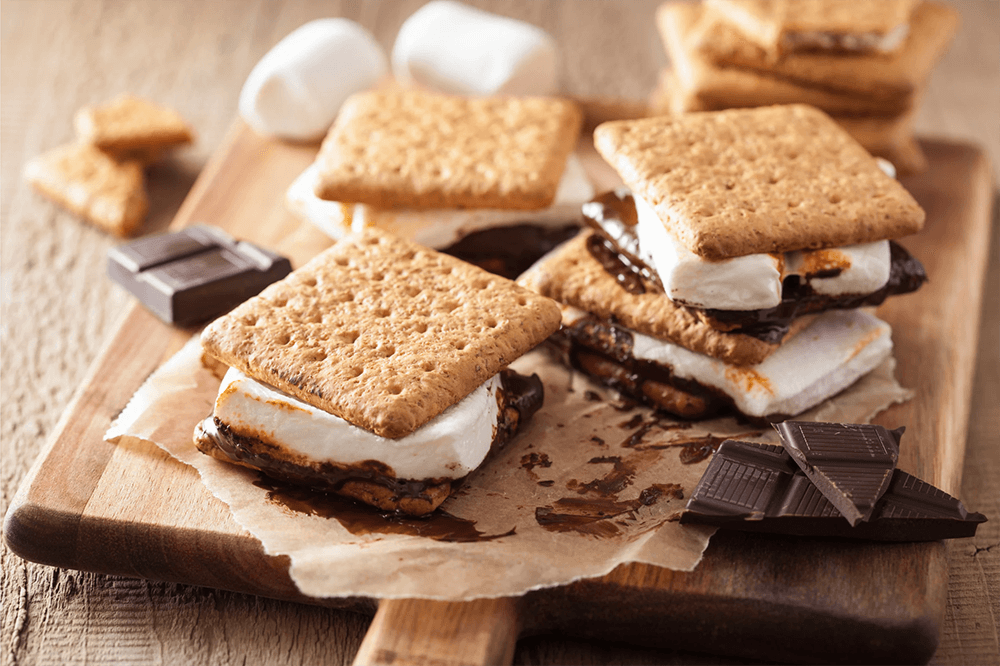 A wooden board with s'mores made of graham crackers, melted chocolate, and toasted marshmallows.
