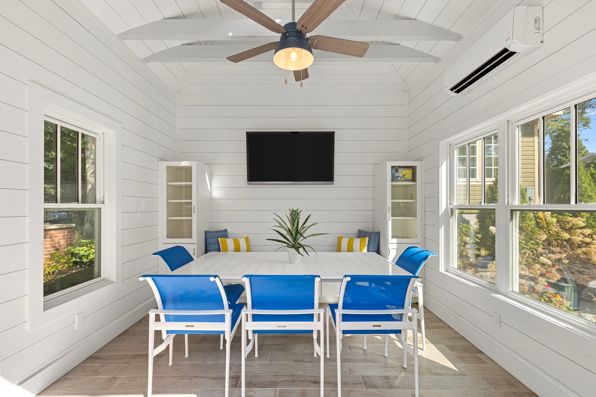 Light-filled sunroom with a white interior, dining table, blue chairs, and a mounted TV.