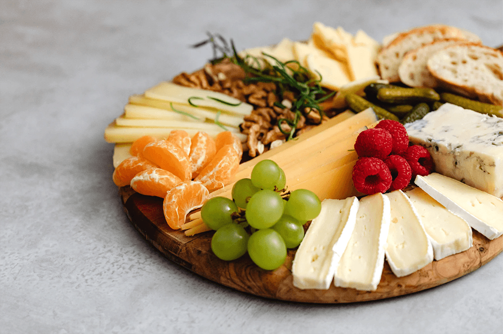 A wooden platter arranged with various cheeses, fruits, nuts, and bread.