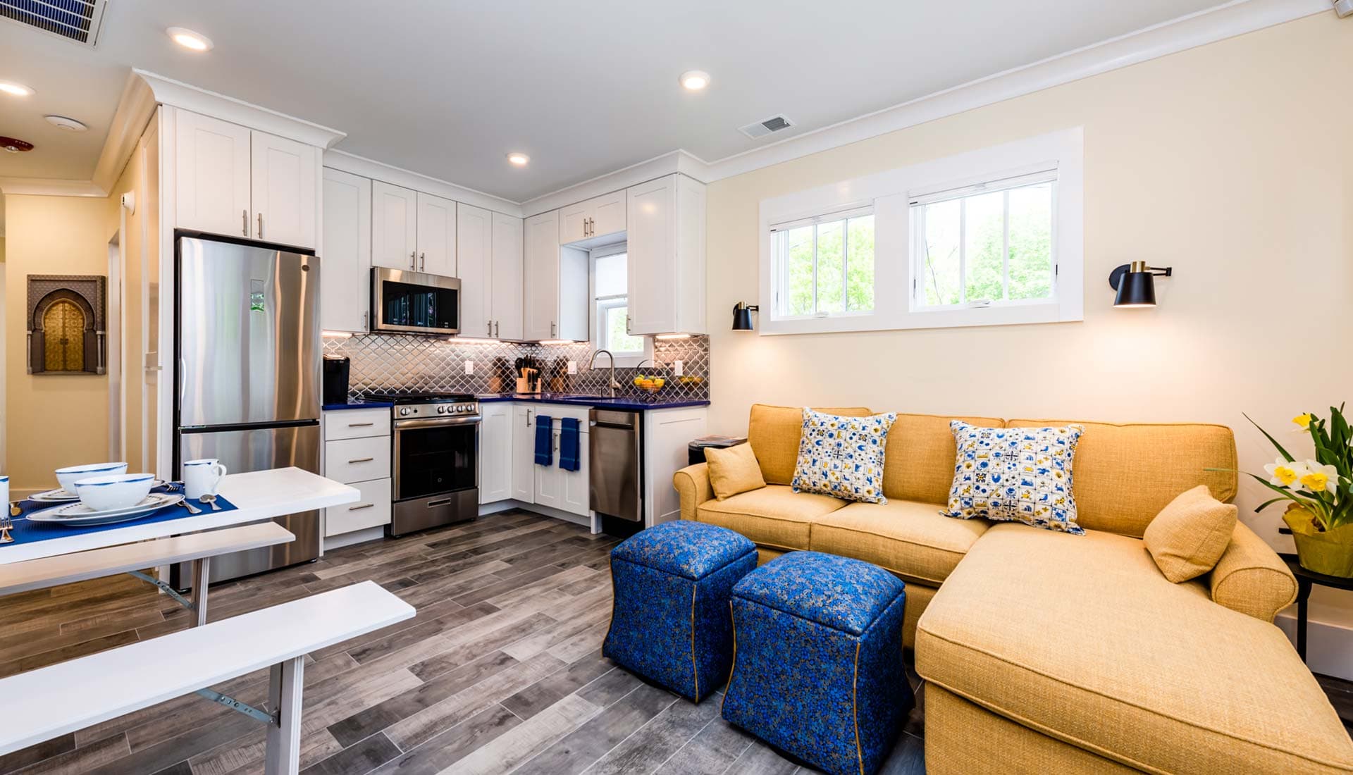 A modern kitchen and living area featuring a sofa with decorative pillows and blue ottomans.