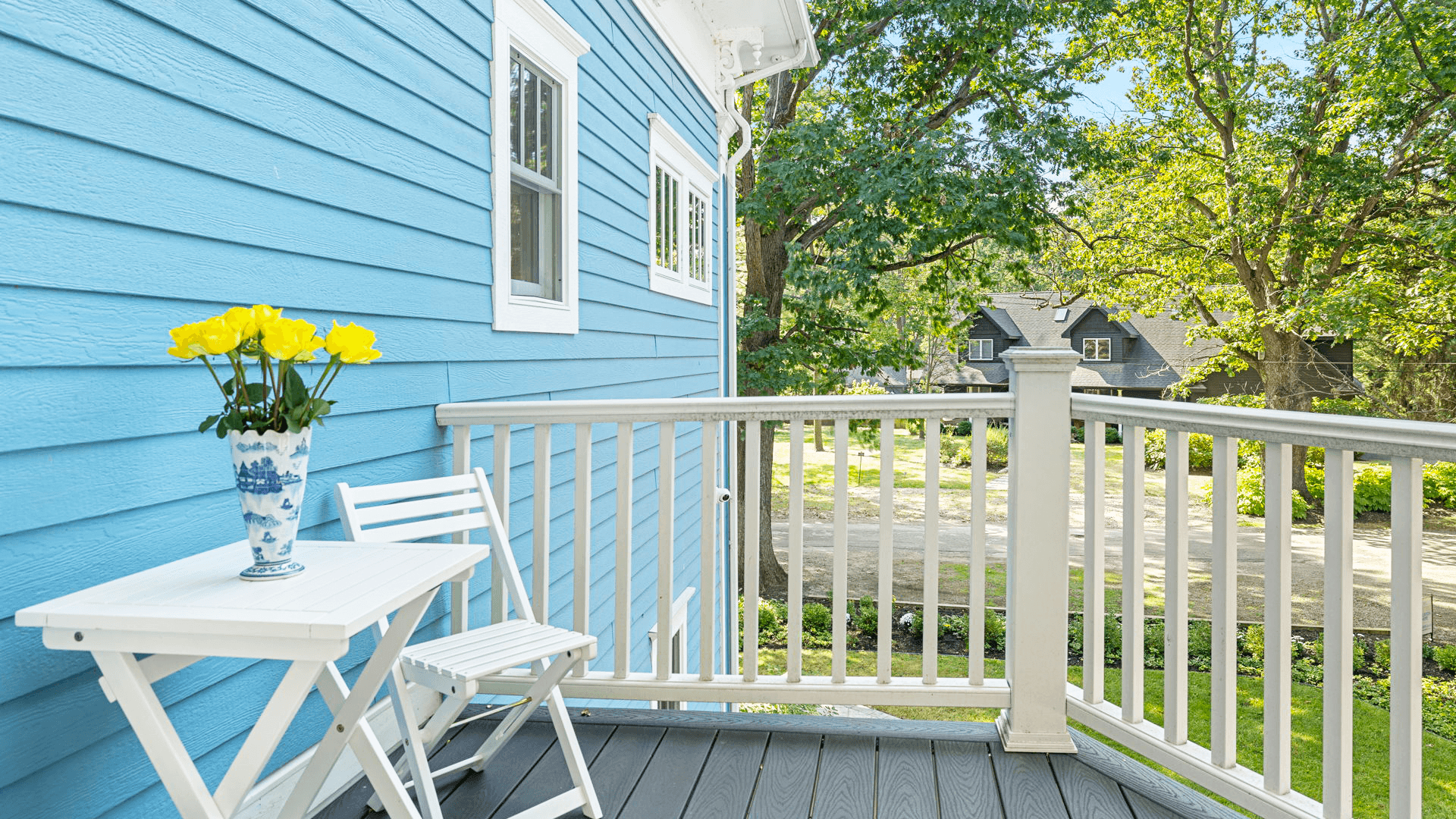 A sunny balcony with a blue wall, white table and chairs, and a vase of yellow flowers.